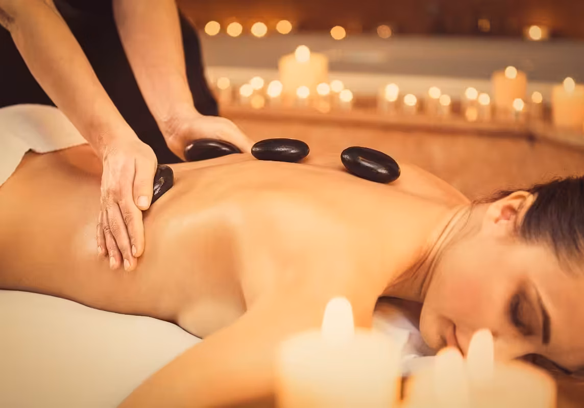 Serene young woman is enjoying massage at beauty salon. Masseuse is touching hot smooth stones to her body